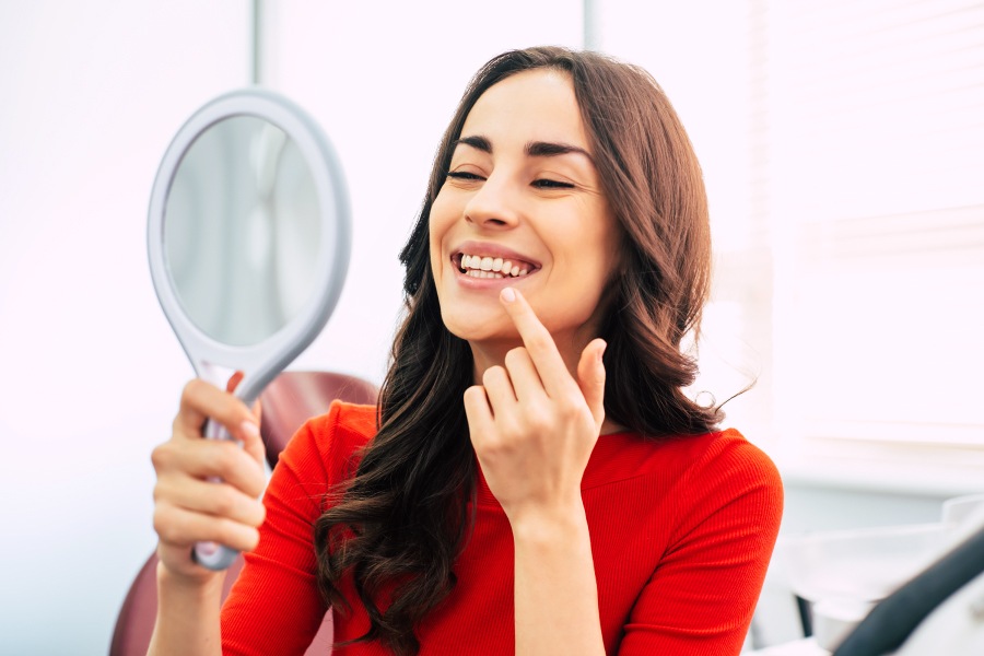 Brunette woman in a red shirt smiles while looking at her teeth in a handheld mirror