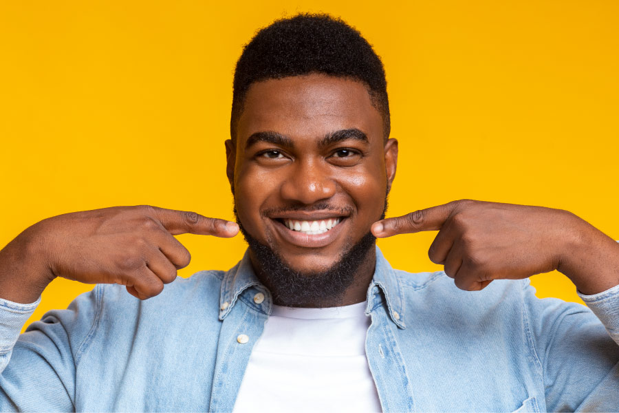 Dark-haired man smiles and points proudly at his beautifully white teeth against a yellow wall