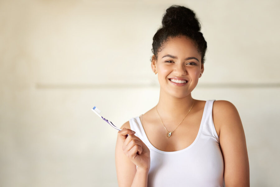 Black young woman in a white tank top smiles while holding her toothbrush in the bathroom before brushing her teeth to prevent cavities