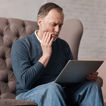 man holding his jaw in pain while looking at a laptop