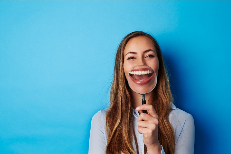 Brunette white woman smiles as she holds up a magnifying glass to her teeth against a blue background
