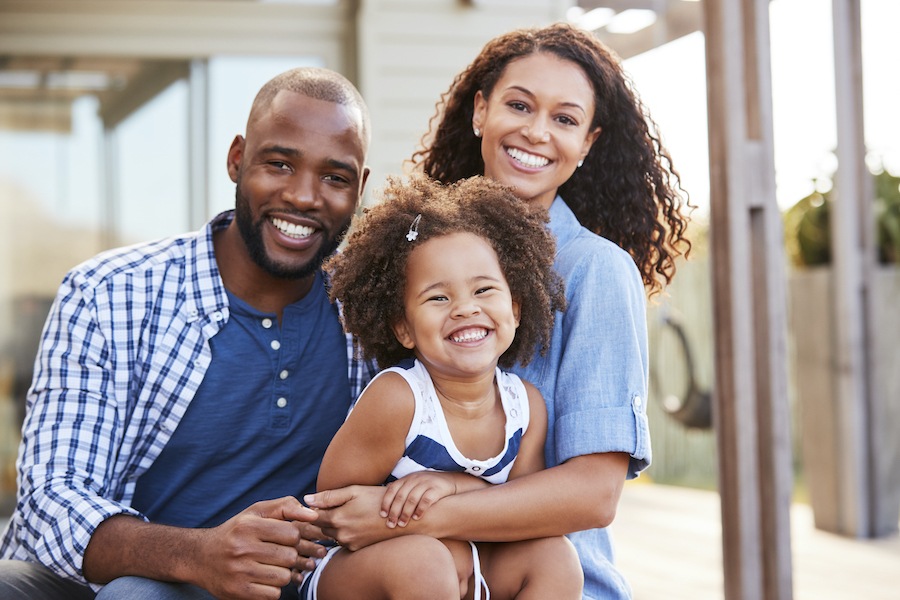 Black mom and dad embrace their adorable young daughter smile smiling outside sitting on a porch