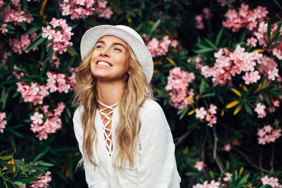 Blonde woman in a white blouse and hat smiles in front of a bush with blooming pink spring flowers