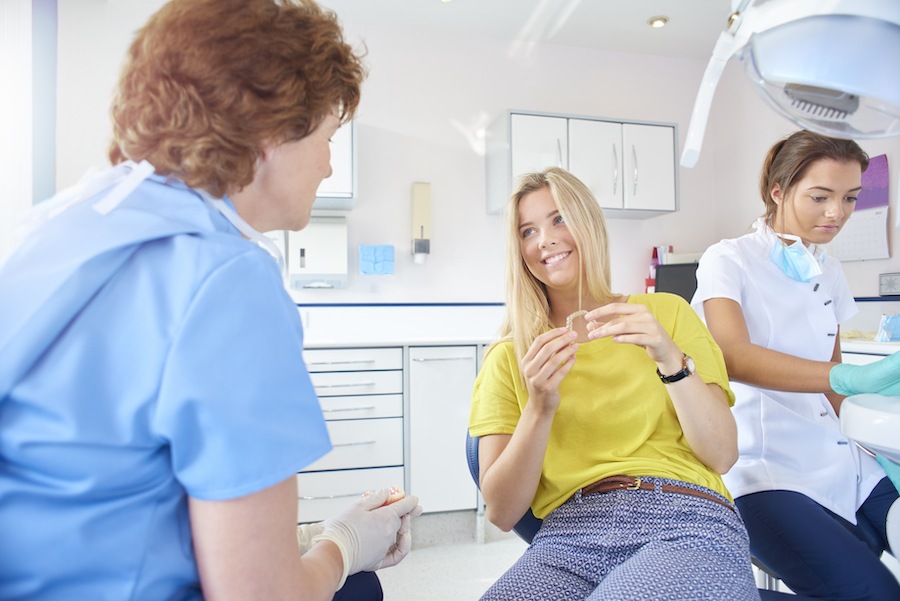 A blonde teenager in a yellow blouse smiles while holding Invisalign clear aligners while sitting in a dental chair