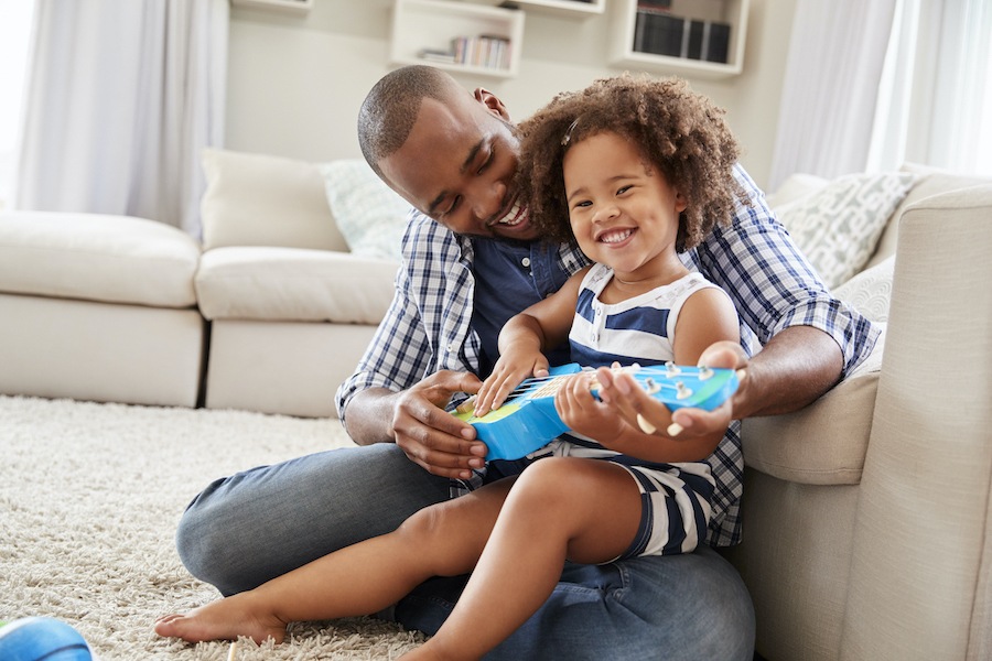 Black dad smiles as he teaches his young daughter how to play the ukulele as she sits on his lap